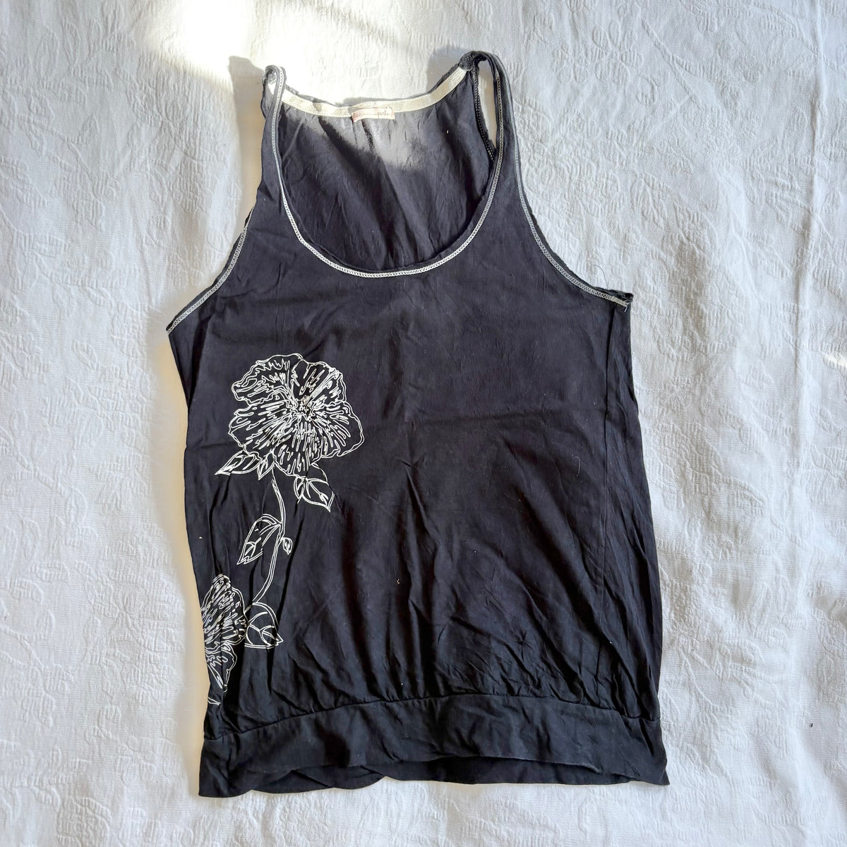 A black sleeveless tank top with a white floral line-art graphic on the front, laid flat on a wrinkled white fabric background.