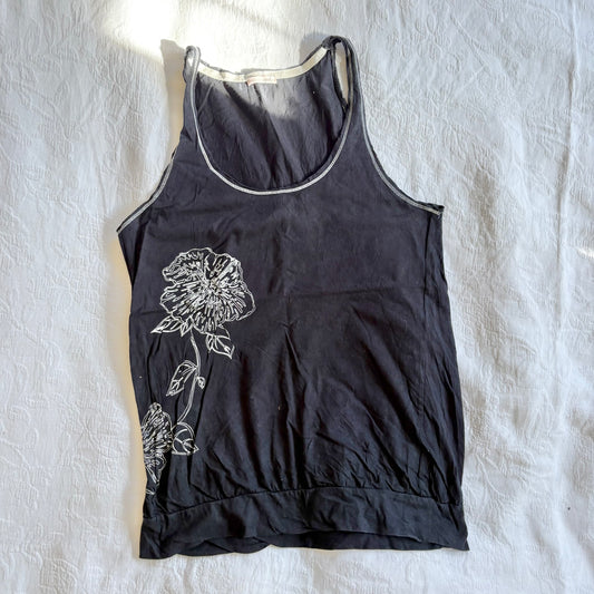 A black sleeveless tank top with a white floral line-art graphic on the front, laid flat on a wrinkled white fabric background.